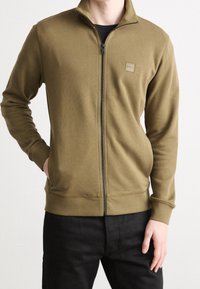 Man som bär en olivgrön zip-up jacka med liten logotyp på bröstet, kombinerad med svarta byxor, står med en hand i fickan.