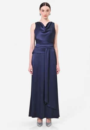 Femme portant une robe de soirée en satin bleu marine sans manches avec un décolleté drapé et des talons hauts argentés, debout sur un fond blanc.