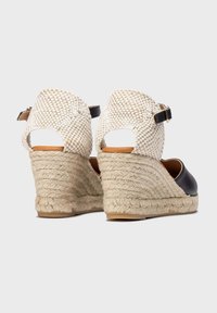 Sandalias de cuña de cuero negro con tacón de tela blanca tejida y correa en el tobillo. Presentan una suela de yute trenzado y un diseño de espalda abierta.