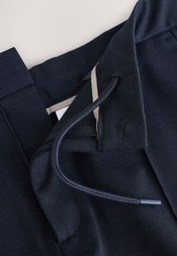 Nahaufnahme von marineblauen Hosen mit geflochtenem Kordelzug, Reißverschluss und knopfbesetztem Bunddetail auf hellem Hintergrund.
