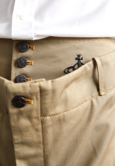 Beiger Baumwollhose mit dunklen Knöpfen, einem orangefarbenen Nähdetail und einem dekorativen schwarzen Metallelement in der vorderen Tasche.