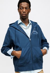 Jauns vīrietis, tērpies zilā kapučjakā ar rāvējslēdzēju pār gaiši zilu kreklu, ar sudraba ķēdītes kaklarotu, rokām ielikts priekšējās kabatās, skatās uz sāniem.