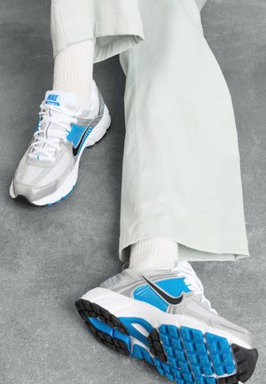 Gray athletic shoes with blue accents and black details. Mesh and synthetic upper, white laces, rubber sole, paired with light green pants.