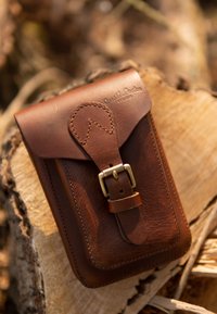 Leren telefoonhoes in diep bruin, met een klep sluiting en een metalen gesp, gegraveerd logo en zichtbare stiksels.