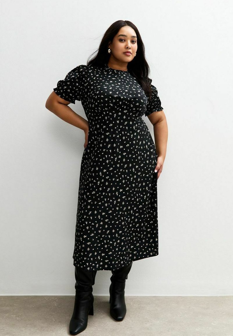 Vestido negro con estampado de flores, con mangas cortas abullonadas y largo hasta la rodilla, que presenta una cintura entallada y una tela suave y texturizada. Combinado con botas negras hasta la rodilla.