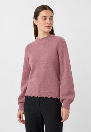 Frau mit glatten braunen Haaren trägt einen rosa strukturierten Pullover mit gewellten Kanten und schwarze Hosen, schaut leicht nach rechts.