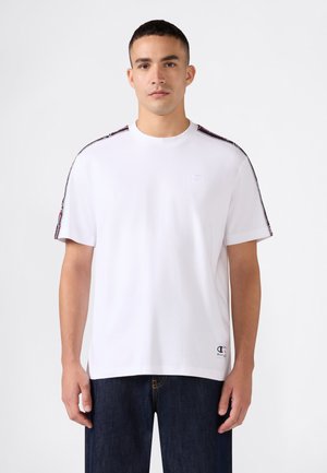 Jonge man met een wit T-shirt met korte mouwen met logostrepen op de schouders en donkerblauwe spijkerbroek, staand tegen een effen achtergrond.