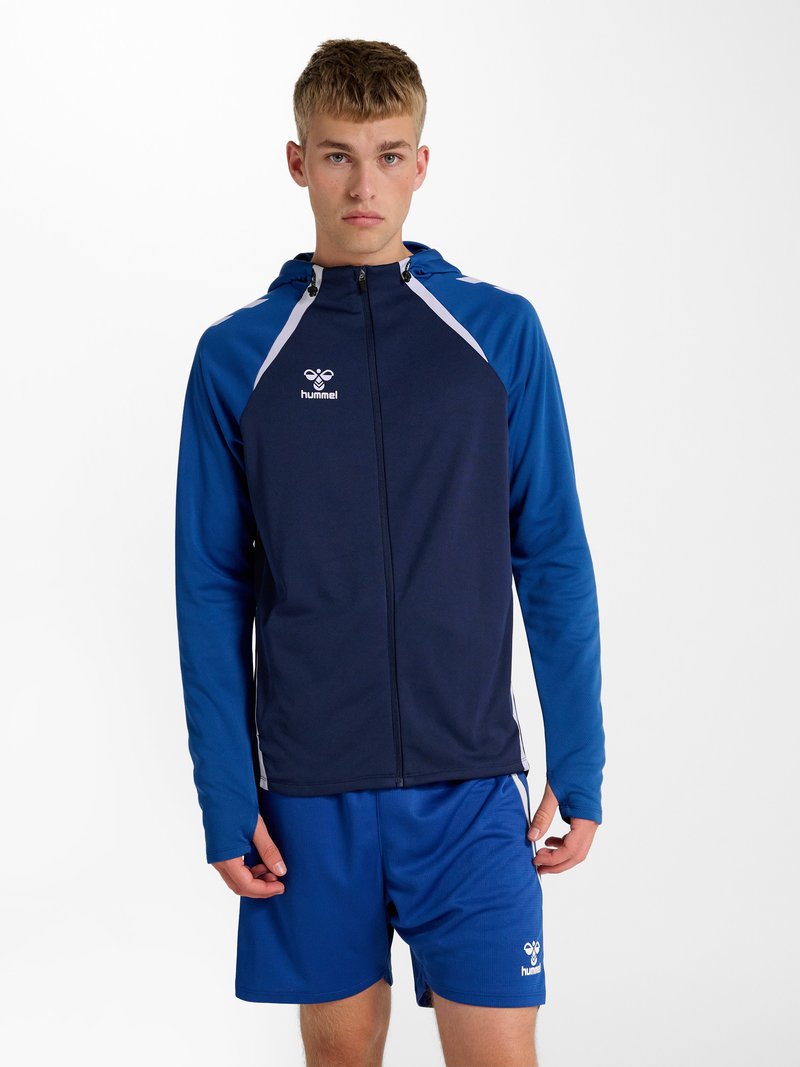Jeune homme portant une veste de sport à capuche bleu marine et bleue avec fermeture éclair et un short bleu assorti, debout devant un fond blanc.