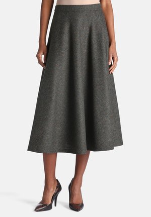 Femme portant une jupe midi en chevrons grise et des chaussures à talons noirs, debout devant un fond uni.