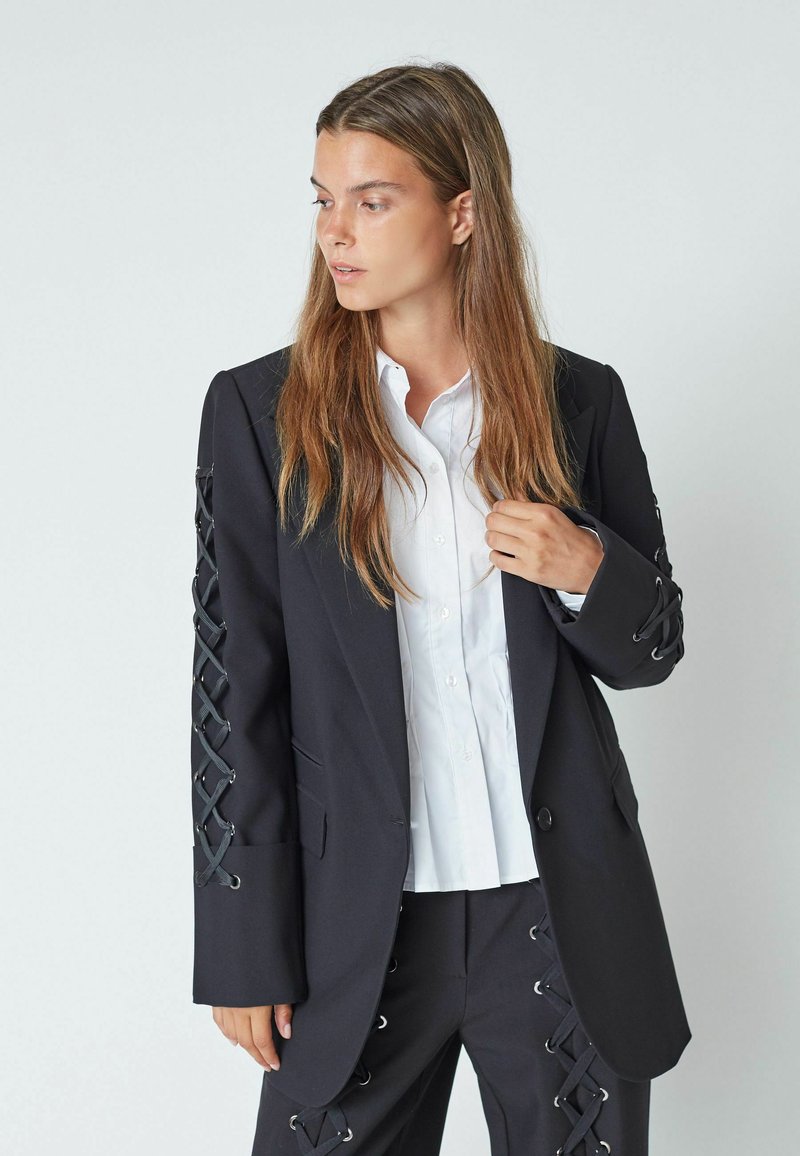 Črn blazer z detajli z vezanjem na rokavih, v kombinaciji z belim srajcami na gumb. Ima prilagojen kroj in mehko teksturo tkanine.