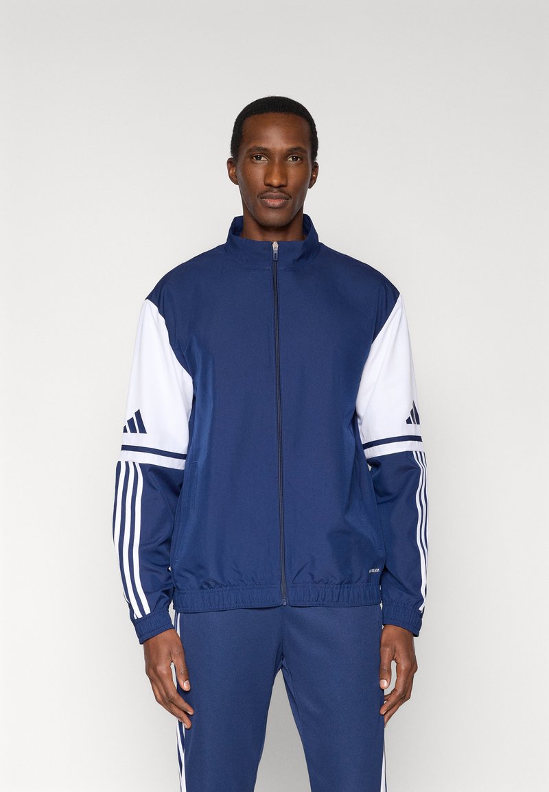 Giacca con zip in blu navy, caratterizzata da maniche bianche, tre strisce su ogni braccio e collo a lupetto. Realizzata in tessuto leggero.