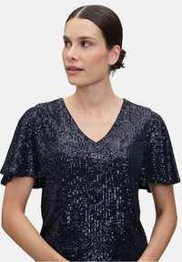 Granatowa bluzka z cekinami z dekoltem w kształcie litery V i krótkimi falbankami. Tkanina charakteryzuje się gęstym rozmieszczeniem cekinów, co nadaje jej teksturowany wygląd.