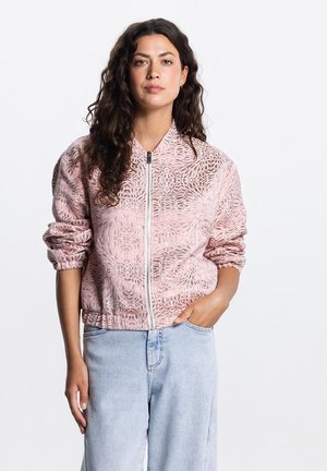 Vrouw met lang krullend haar, gekleed in een roze geribbelde jas met rits en lichtblauwe hoge taille jeans, met één hand in de zak.