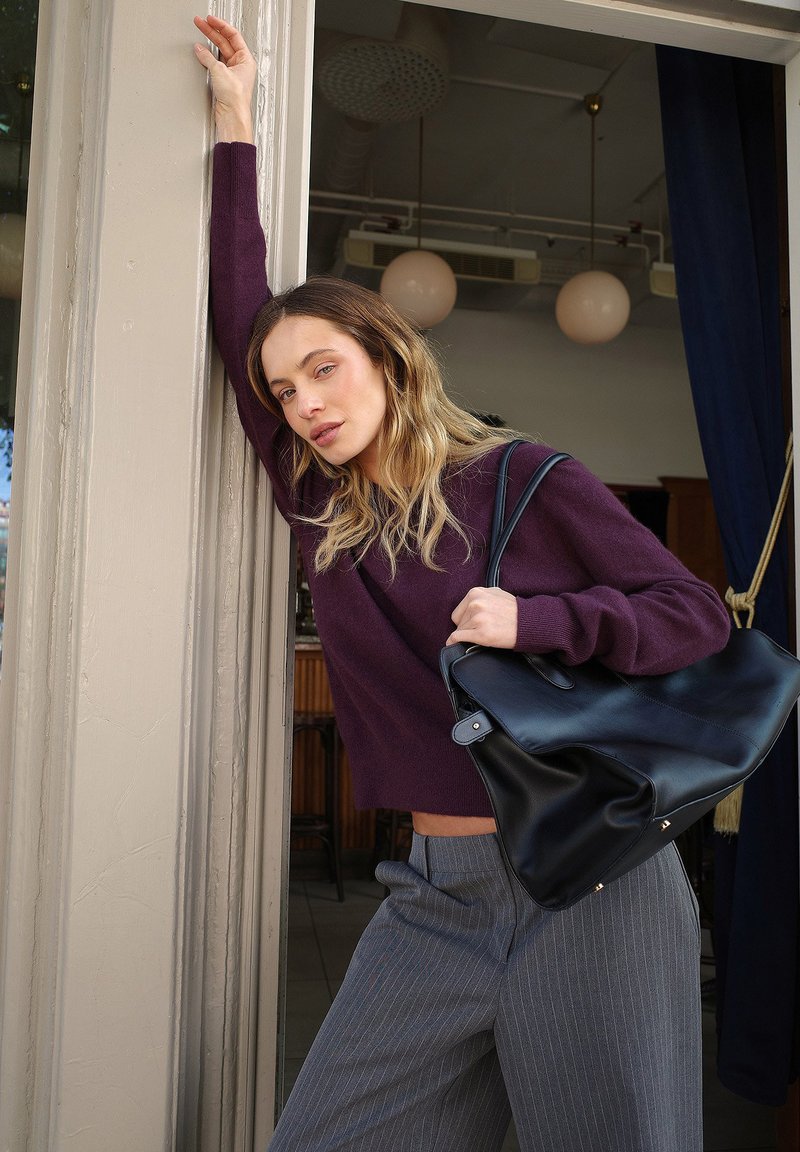 Juodas odinis tote krepšys su lygiu paviršium ir metaliniais akcentais; derinamas su giliu purpuriniu megztiniu ir pilkomis dryžuotomis kelnėmis.