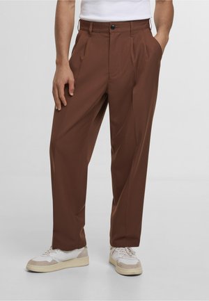 Mann iført høytlivs brune bukser med folder og sidelommer, og hvite joggesko, stående på en ensfarget grå bakgrunn.