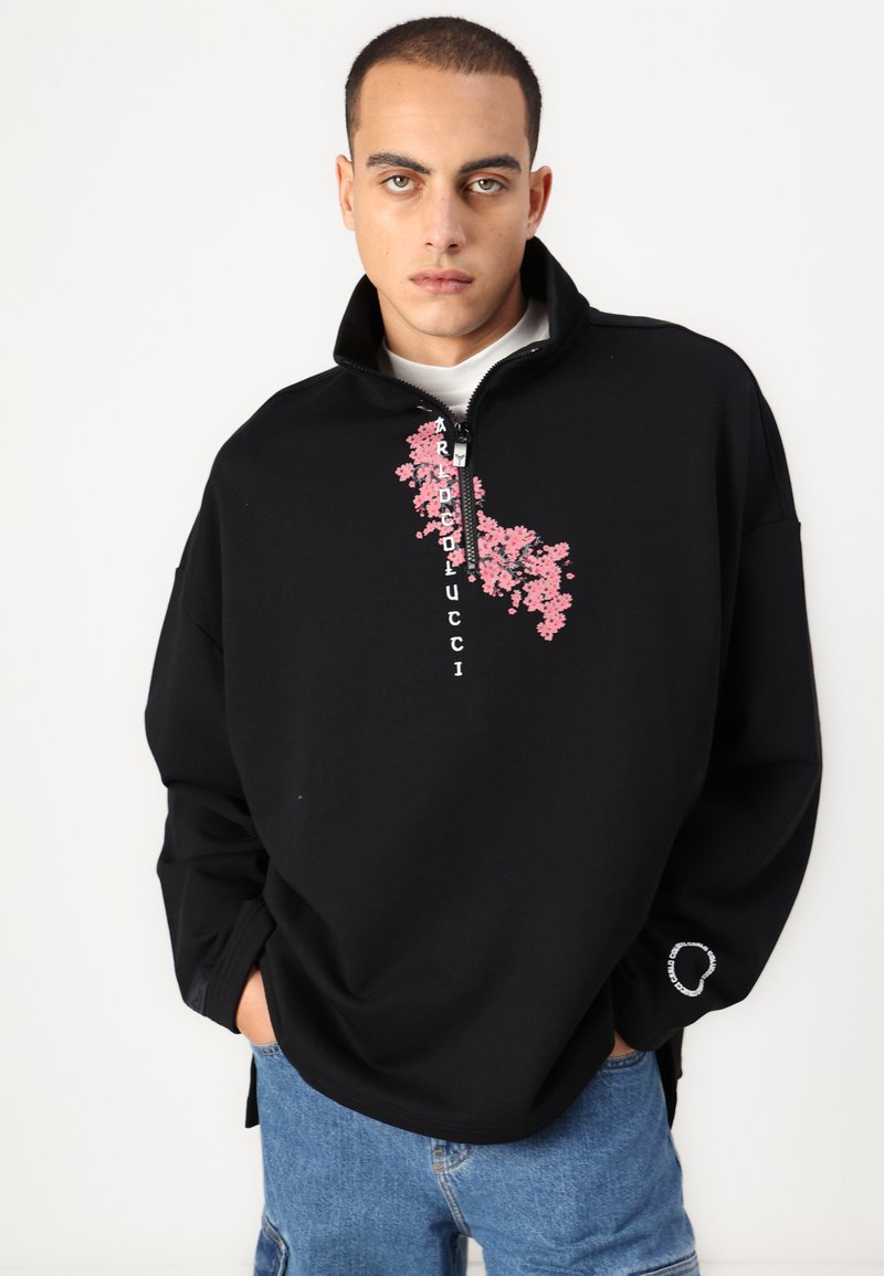 Zwarte fleece sweatshirt met een halflange ritskraag, voorzien van een bloemenprint in het roze en een tekstontwerp op de voorkant. Losse pasvorm met zijsplitten.