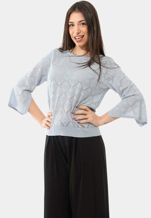 Giovane donna sorridente con le mani sui fianchi, indossa un maglione azzurro chiaro con motivo e maniche svasate, abbinato a pantaloni neri a gamba larga.