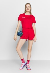 Rød sportsoutfit med kortærmet top og shorts, der har hvid kant. Modellen holder en blå og grøn håndbold og har farverige sportsko og sokker på.