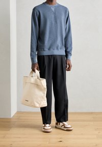 Blauwe crew-neck sweatshirt, zwarte wijde broek, beige schoudertas en bruin gestreepte sneakers met witte zool en veters. Eenvoudige, casual outfit.