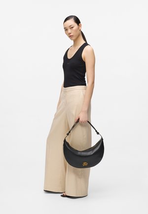 Kobieta w czarnej bluzce bez rękawów i beżowych szerokich spodniach, trzymająca czarną torebkę w kształcie półksiężyca z złotym logo, stojąca na jednolitym tle.