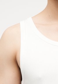 Nærbilde av en persons øvre kropp som har på seg en hvit ribbestrikket singlet mot en ensfarget lys bakgrunn.