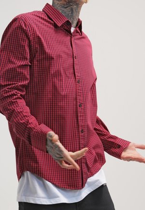 Hombre con tatuajes en el cuello y las manos, lleva una camisa roja y negra a cuadros sobre una camiseta blanca con los brazos ligeramente extendidos.