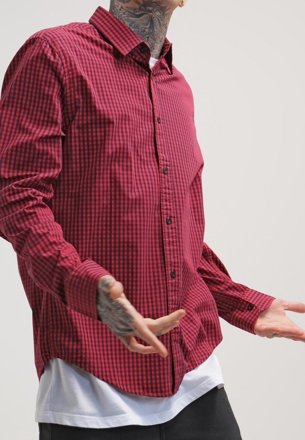 Homme avec des tatouages au cou et aux mains, portant une chemise à carreaux rouge et noir par-dessus un t-shirt blanc, les bras légèrement étendus.