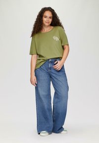 Olivegrünes, übergroßes T-Shirt mit Textgrafik; kombiniert mit weit geschnittenen blauen Denim-Jeans. Das Model steht vor einem neutralen Hintergrund.