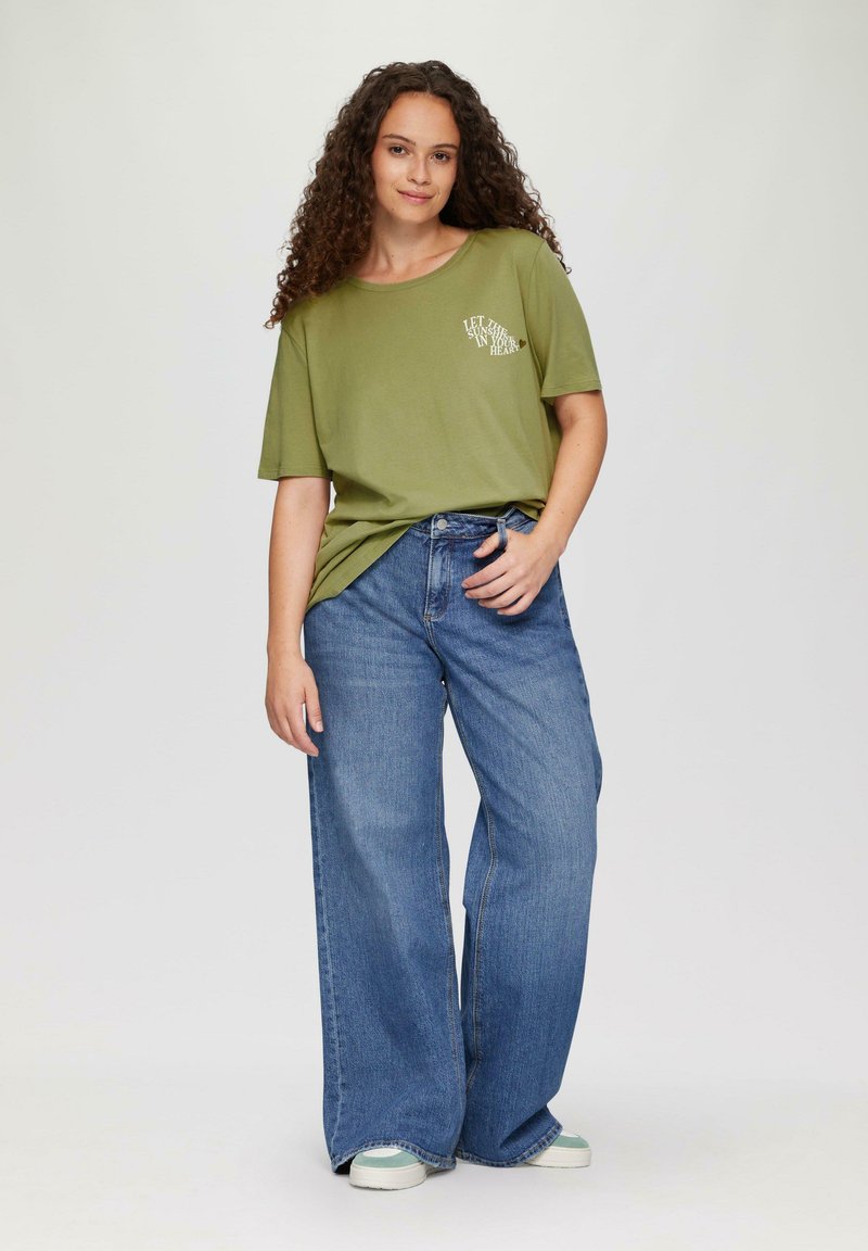 Olivegrünes, übergroßes T-Shirt mit Textgrafik; kombiniert mit weit geschnittenen blauen Denim-Jeans. Das Model steht vor einem neutralen Hintergrund.