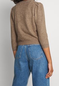 Hnědý pletený svetr s tříčtvrtečními rukávy a sklady na ramenou, doplněný světle modrými džínami s vysokým pasem.