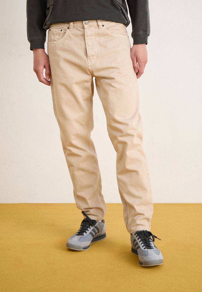 Jean en coton beige avec une coupe droite, présentant une fermeture à bouton et un design à cinq poches, associé à des chaussures de sport grises.