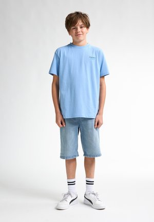 Hellblaues Baumwoll-T-Shirt, lockere Passform. Helle Jeansshorts mit geradem Schnitt. Weiße Sneakers mit schwarzen Akzenten, knielange gestreifte Socken.