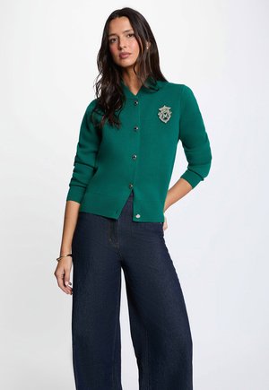 Donna con lunghi capelli scuri che indossa un cardigan verde con bottoni e stemma e jeans scuri a gamba larga su uno sfondo chiaro uniforme.