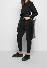 Černý neformální outfit skládající se z dlouhého rukávu s kapucí, kalhot s tkaničkou v pase a černých dřeváků. Je nošena dlouhá černá taška.