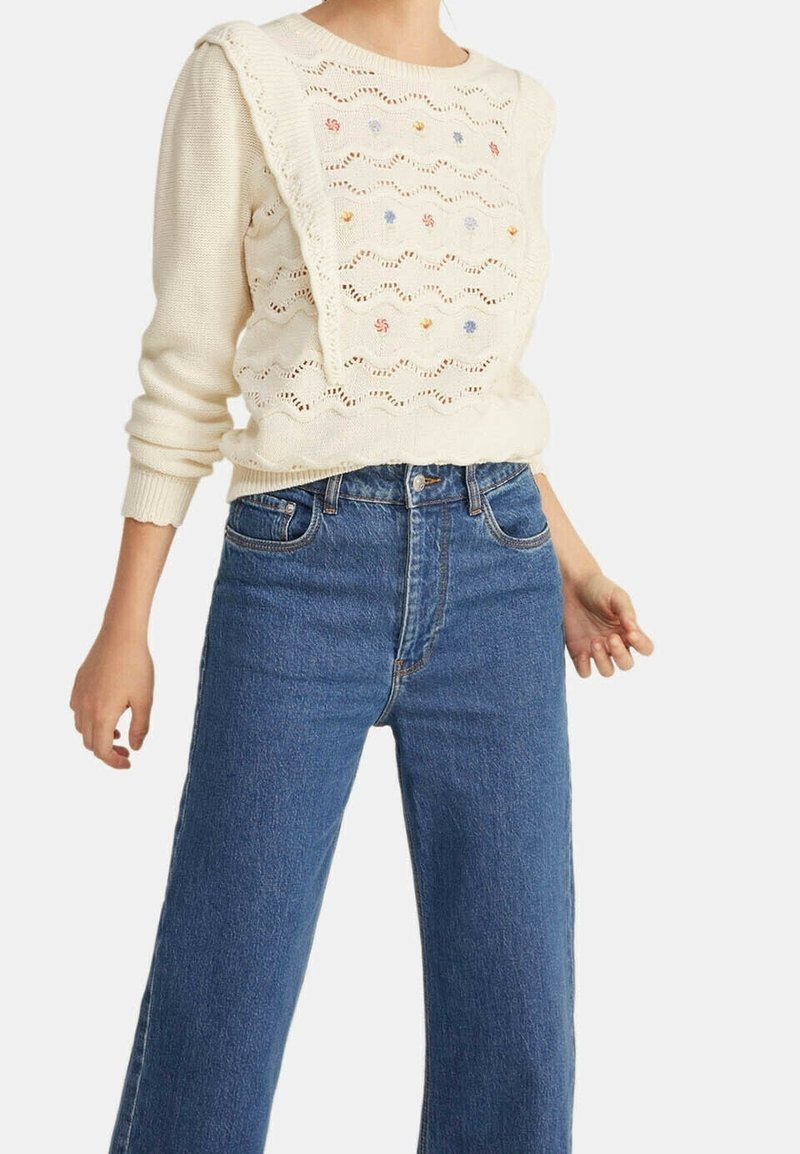 Gruba dzianinowa sweter z wzorem ażurowym, kolorowymi haftami i złożonym panelem, zestawiony z niebieskimi dżinsami o wysokim stanie.