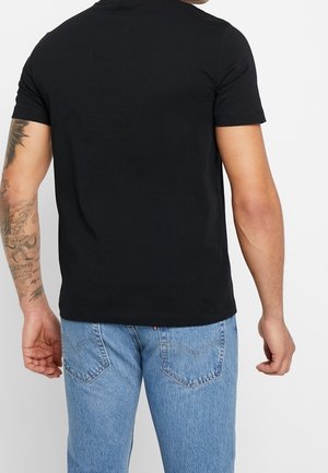 Homme portant un t-shirt noir à manches courtes uni et un jean bleu clair, montrant son bras gauche tatoué, se tenant devant un fond blanc.