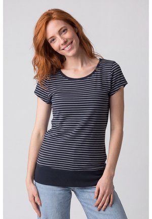 Junge Frau mit roten Haaren lächelt, trägt ein schwarz-weiß gestreiftes kurzärmeliges Shirt und hellblaue Jeans, steht vor einem schlichten Hintergrund.