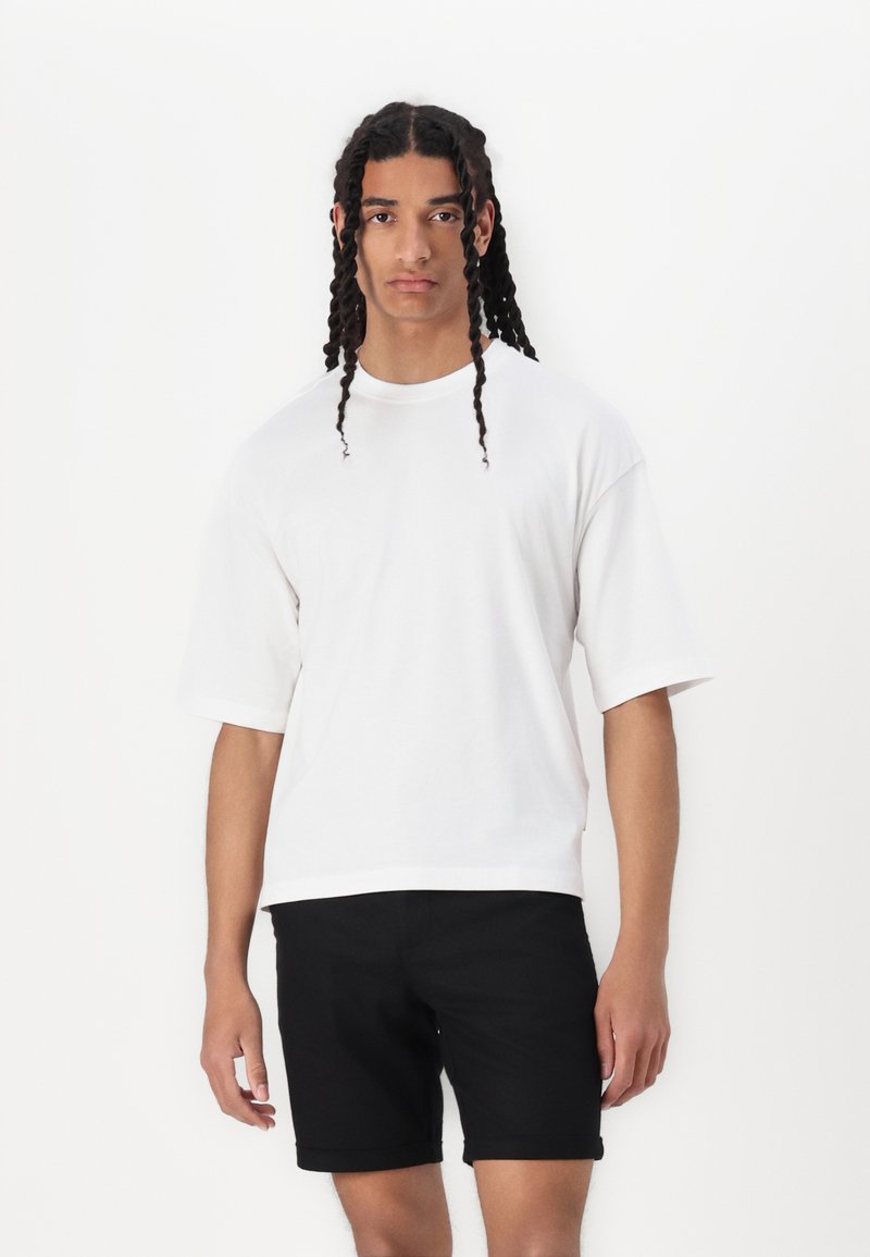 White oversized t-shirt with short sleeves paired with black shorts. Simple, clean design; smooth texture with a relaxed fit.