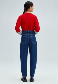 Camisa roja de gran tamaño y manga larga con puños a rayas en blanco y negro, combinada con pantalones vaqueros de tiro alto en azul y botas negras con tobillo.