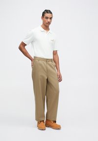 Witte poloshirt met een kraag en een klein logo, gecombineerd met tan brede pantalons en donker tan werkschoenen. Glad textuur door het geheel.