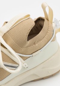 Zapatilla de tela beige con diseño sin cordones, acentos blancos, acabado texturizado y collarín acolchado en el tobillo con cordones elásticos.