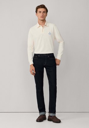 Helle, langärmlige Poloshirt mit Kragen und blauem Logo, kombiniert mit dunklen Denim-Jeans und braunen Schuhen. Neutraler Hintergrund.