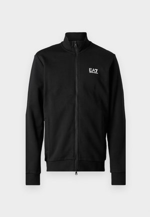 Svart zip-up jakke laget av glatt stoff, med stående krage, tettsittende ermer og en liten hvit logo på brystet.