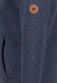Sudadera de algodón azul marino con patrones salpicados en rosa y un parche redondo de cuero marrón con un diseño de corazón en la manga.