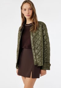 Veste matelassée vert olive avec une texture brillante, dotée de fermetures à boutons-pression et d'un col montant. Portée sur un haut et une jupe bruns.
