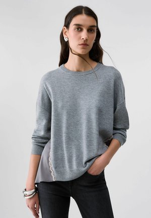 Donna con capelli lunghi e scuri indossa un maglione grigio sopra pantaloni neri, orecchini a forma di cuore in argento e braccialetti d'argento, in piedi con una mano in tasca.