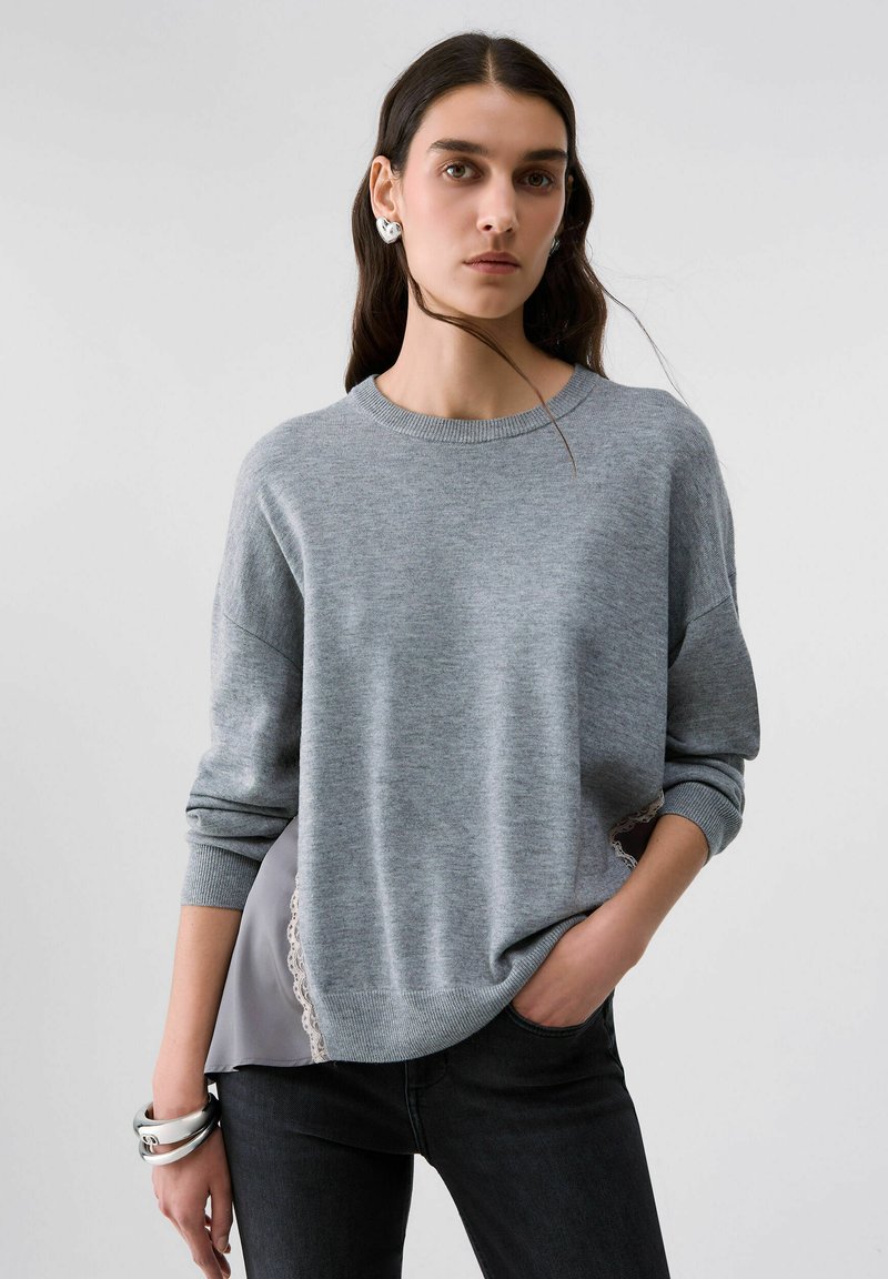 Femme aux longs cheveux foncés porte un pull gris sur un pantalon noir, des boucles d'oreilles en forme de cœur en argent, et des bracelets en argent, debout avec une main dans la poche.