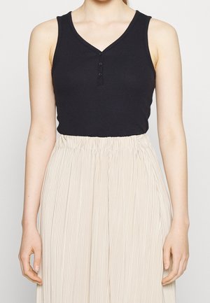 Femme portant un top noir sans manches avec boutons et col en V, rentré dans une jupe beige plissée à taille haute.