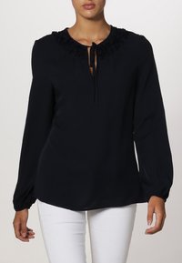 Černá blůza s dlouhým rukávem, s texturovaným volánkovým výstřihem a detailem vázání. Hladká tkanina s uvolněným střihem, kombinováno s bílými kalhotami.