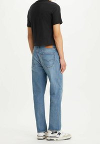 Jeans de mezclilla azul con corte recto, que presentan una etiqueta de cuero en la parte trasera y costuras visibles. Combinados con una camiseta negra y zapatillas.